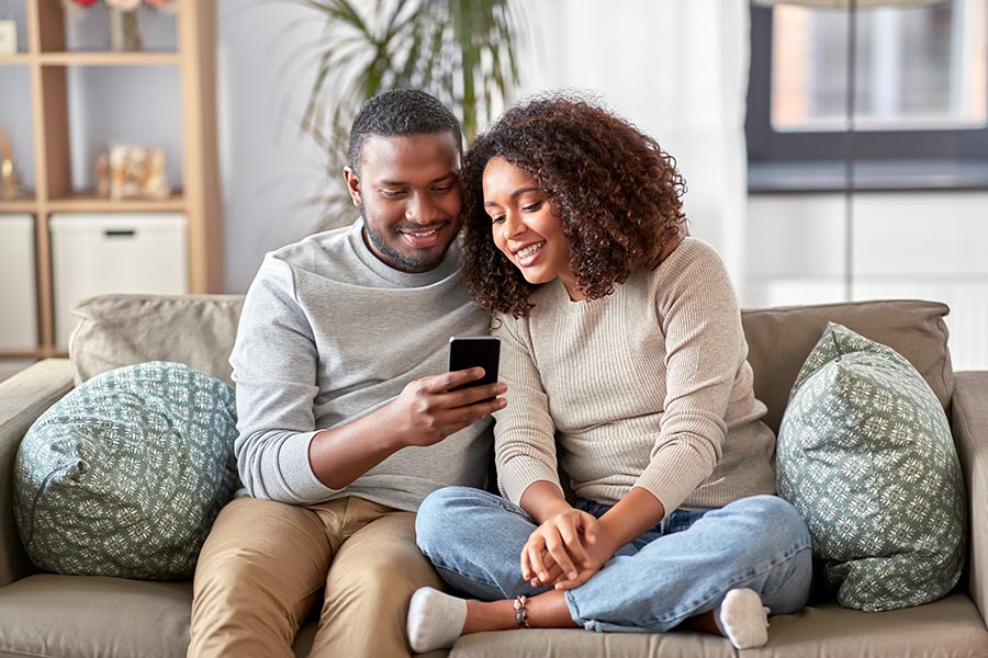 Contact Us - Young Couple Sits Close on Their Sofa, Looking at Husband's Smartphone, Potted Plants and Bookshelves Behind Them