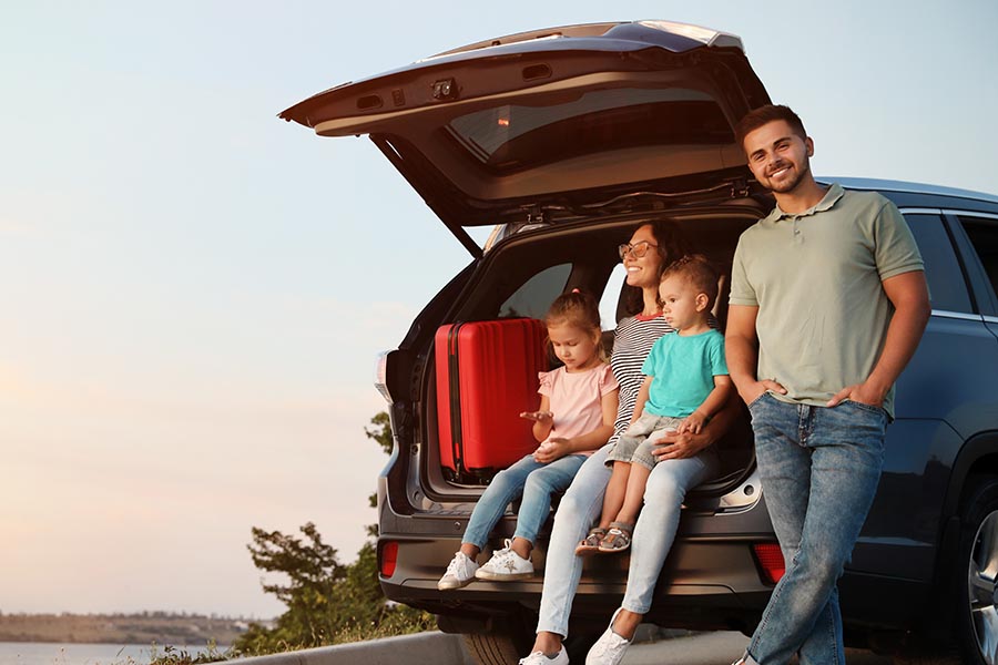 Personal Insurance - Young Family Sits in the Tailgate of Their Car, Packed for a Road Trip, Stopping to Enjoy a Lake View
