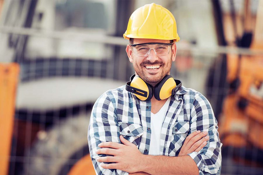 Contractor Insurance - Portrait of a Smiling Young Construction Worker in a Hard Hat with Arms Crossed Standing in Front Construction Equipment at a Construction Site