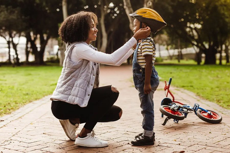 Group Voluntary Supplemental Life Insurance - Mother Placing a Protective Helmet on Her Son Before Riding a Bike in the Park on a Sunny Day